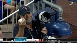 Man holding baseball and camera equipment at stadium during game broadcast.