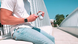 Person in white shirt holding smartphone while sitting outdoors against corrugated metal buildings.