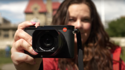 Woman holding a Leica camera toward the viewer, with a historic building blurred in the background.
