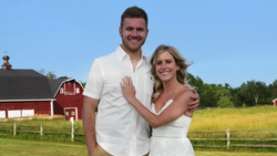 Couple in white clothing posing together in front of a red barn and green pasture.