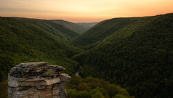 Weathered rock outcrop overlooking a forested valley at sunset, with layered mountains receding into golden light.
