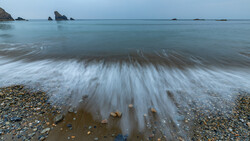 Long-exposure photograph of waves washing across a pebble beach at dusk, with rocky outcrops visible in the calm sea beyond.