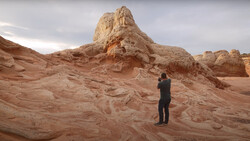Photographer in dark clothing standing on layered sandstone formations, framing a tall rock pinnacle with camera.
