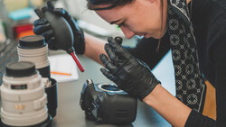 Photographer in black gloves examining a camera lens at a workbench with equipment and lenses nearby.
