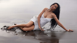 Woman in white clothing reclining on wet sand near shallow water at a beach.