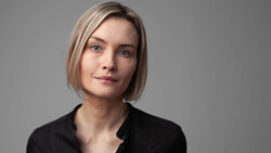Woman with shoulder-length blonde hair wearing a black shirt against a neutral gray background.