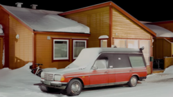 Red and white hearse parked in front of wooden cabin structures in heavy snow.