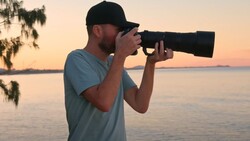 Male photographer holding a telephoto lens to his eye during golden hour near water.
