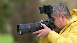 Photographer in yellow jacket holding camera with telephoto lens and electronic viewfinder.
