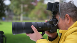 Photographer in yellow jacket holding Nikon camera with telephoto lens aimed at outdoor subject.