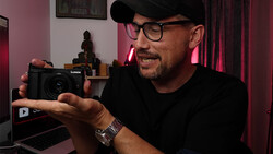 Photographer holding a Lumix compact camera while reviewing footage on a laptop in a studio setting.
