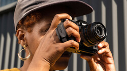 Photographer holding a rangefinder camera up to their face, wearing a cap and hoop earrings against a striped metal background.