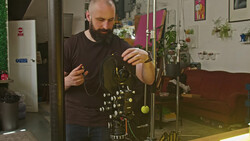 Bearded man examining a complex mechanical device with brass components and hanging weights in a studio setting.