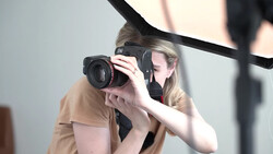 Photographer holding a mirrorless camera with telephoto lens to her eye while shooting in a studio setting.