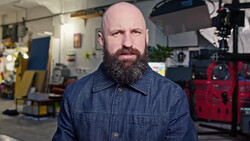 Man with full beard wearing denim shirt in workshop studio setting.