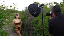 Photographer shooting a woman in bronze-toned clothing outdoors using a large parabolic softbox reflector.