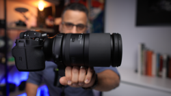 Photographer holding a mirrorless camera with telephoto lens extended toward the camera.