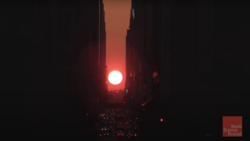 Bright red sun framed by tall buildings in a narrow urban canyon at dusk.