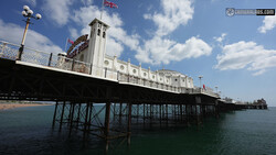 Victorian-era seaside pier with white ornamental structure and iron support columns extending into calm turquoise water under blue sky.