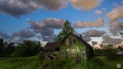 Abandoned wooden house surrounded by overgrown vegetation under a dramatic cloudy sky.