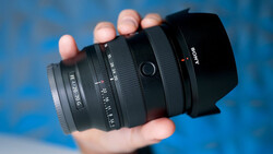 Hand holding a Sony camera lens with black barrel and hood against blue background.