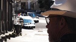 Man in cowboy hat and sunglasses looking down a sunlit alley lined with buildings and parked vehicles.