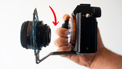 Camera lens mounted on a mirrorless camera body held in a hand, with a red arrow indicating the lens attachment point.