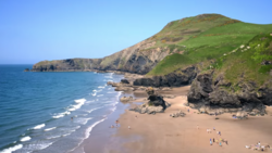 Sandy beach backed by steep green cliffs with rocky outcrops and ocean waves rolling toward shore.