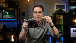 Man in glasses holding a Fujifilm mirrorless camera while speaking to the camera.