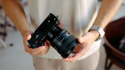 Person holding a black rangefinder camera with attached zoom lens against a light-colored shirt.