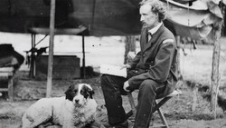 Man in suit seated outdoors holding a document, with a seated dog beside him.