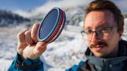 Photographer holding colorful optical filters up to camera lens outdoors in snowy mountain setting.
