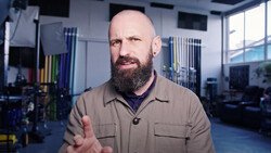 Bald man with full beard wearing tan work shirt gesturing while speaking in a gym setting.
