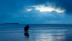 Silhouetted figure crouching on wet beach sand at twilight beneath dramatic cloud formations.