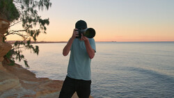 Photographer holding binoculars to eyes while standing on a beach at sunset.