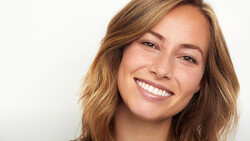 Woman with wavy brown hair smiling directly at camera against white background.