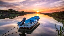 Blue fishing boat anchored in still water at sunrise with golden sky and marsh grasses.