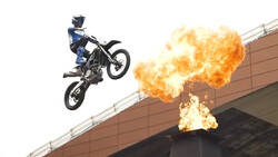 Motocross rider airborne on dirt bike beside massive explosion of flames.