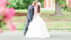Bride and groom kiss outdoors during wedding ceremony, surrounded by pink flowering shrubs and trees.