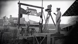 Film crew operating a movie camera on a platform overlooking a European city during early cinema production.