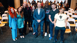 Group of eight people posing together at an indoor event with wooden chairs visible in the background.