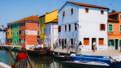 Brightly colored buildings reflected in a canal with moored boats in the foreground.