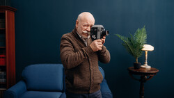 Bald man in brown cardigan holding vintage film camera, standing against dark teal wall.