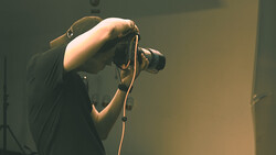 Photographer holding a DSLR camera up to their face, composing a shot in warm studio lighting.