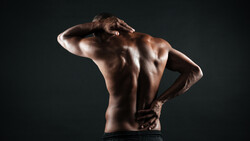 Shirtless man from behind holding lower back with both hands, displaying muscular definition against dark background.