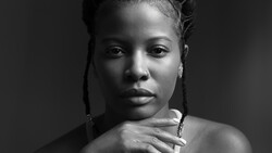 Black and white portrait of a woman with braided hair, looking downward with her hand near her shoulder.