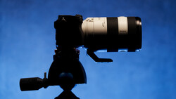 Silhouetted camera on tripod with telephoto lens mounted against a clear blue sky.