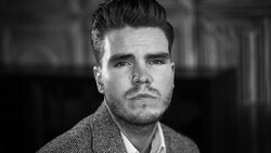 Black and white portrait of a man with styled dark hair and facial hair, wearing a herringbone pattern jacket.