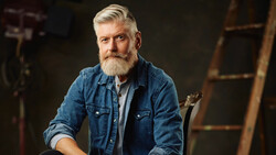 Bearded man in denim shirt posed against dark industrial background.