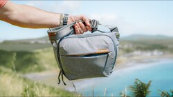 Person holding a gray camera bag against a coastal landscape with turquoise water and green hills.